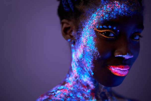 black lady with neon make-up body art posing looking at camera, isolated on purple - Foto, Imagen