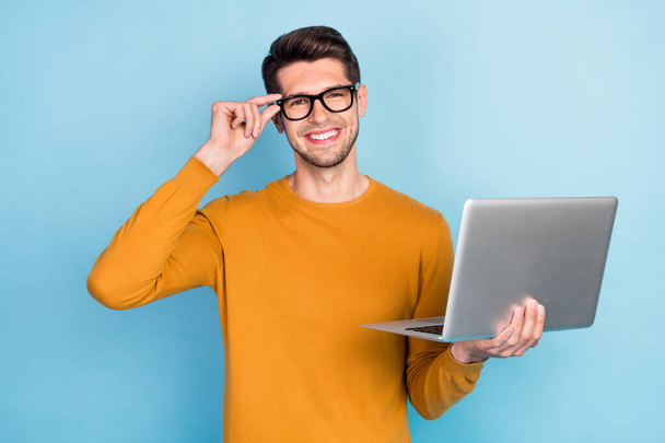 Photo of pretty attractive young man wear yellow sweater arm spectacles smiling holding modern gadget isolated blue color background - Foto, afbeelding