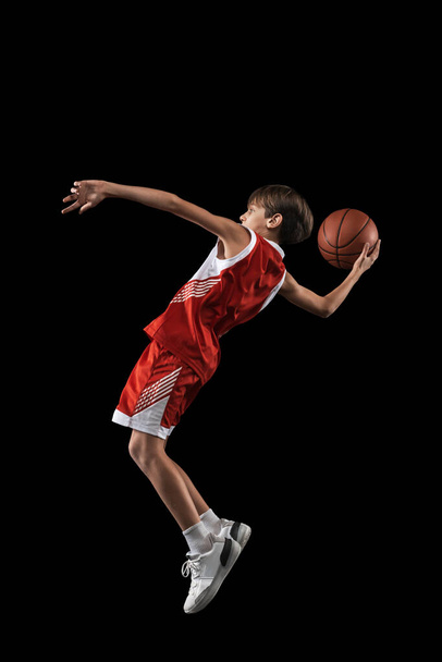 Full-length portrait of teen boy, basketball player in motion, training, playing isolated over black background. Side view - Foto, afbeelding