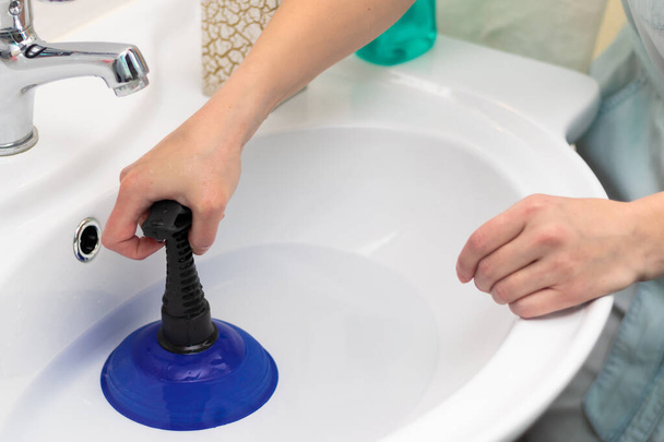 Μια γυναίκα καθαρότερη σε ένα φωτεινό μπάνιο καθαρίζει ένα μπλοκάρισμα στο νεροχύτη με ένα έμβολο. Επιλεκτική εστίαση. Κοντινό πλάνο - Φωτογραφία, εικόνα