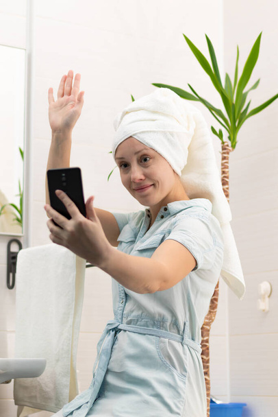 Młoda kobieta z białym ręcznikiem na głowie w jasnej łazience komunikuje się ze smartfonem z przyjacielem w sieciach społecznościowych. Refleksja w lustrze. Wybiórcze skupienie. Portret - Zdjęcie, obraz