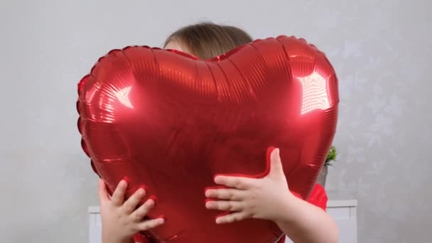 little cute girl in a red dress hides behind a red heart shaped balloon. valentines day concept - Video