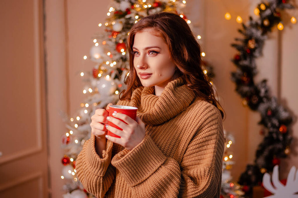 Close-up portrait of an attractive young woman wearing a warm sweater with a red mug in her hands on the background of a Christmas tree. The girl drinks a drink in a cozy New Year's interior. - 写真・画像