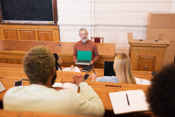 Profesor sosteniendo portátil con pantalla verde cerca de estudiantes multiétnicos en el auditorio de la universidad  - Foto, imagen