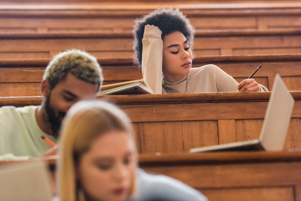 African american student holding pen near laptop and blurred friends in university  - Fotografie, Obrázek