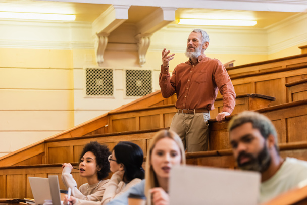 Mature enseignant parler près flou interracial étudiants avec des ordinateurs portables à l'université  - Photo, image