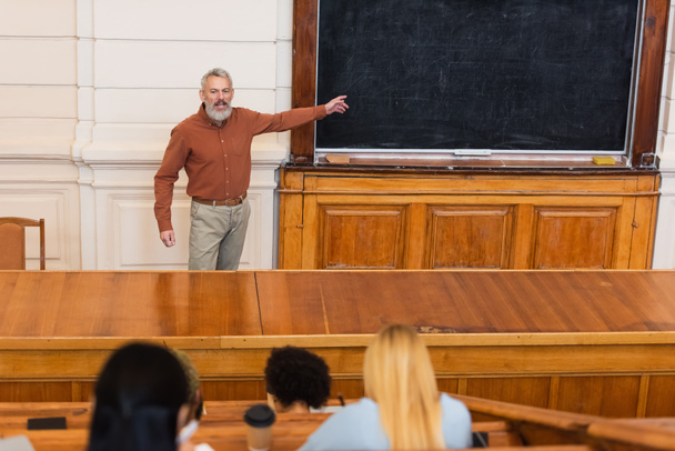 Profesor apuntando a pizarra cerca borrosa estudiantes interracial en la universidad  - Foto, Imagen