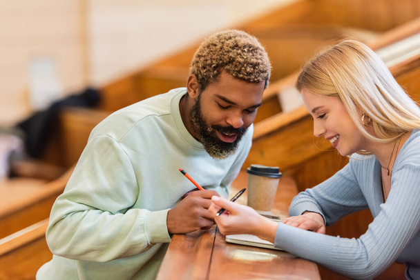 Positive interracial students looking at notebook near coffee to go in university  - Photo, Image