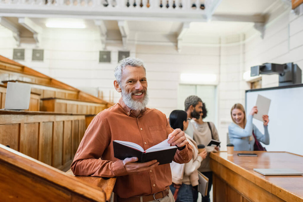 Profesora madura positiva sosteniendo cuaderno cerca de estudiantes borrosos en auditorio universitario  - Foto, Imagen