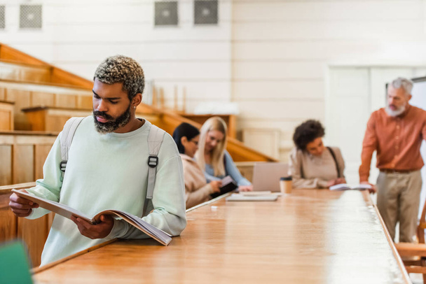 Étudiant afro-américain tenant un cahier près d'amis flous et professeur dans l'auditorium universitaire  - Photo, image