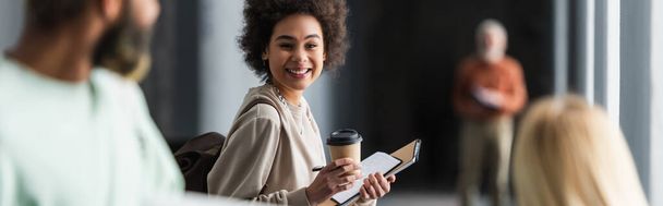 Cheerful african american student holding coffee to go and notebooks near blurred friends in university, banner  - Foto, imagen