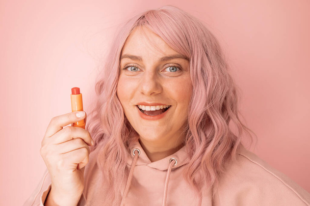 Charmante vrouw houden lippenstift pomade voor te bereiden op datum geïsoleerd over pastel roze achtergrond - Foto, afbeelding