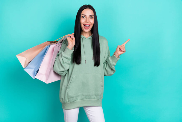 Photo of hooray young brunette lady indicate promo hold bags wear green pullover isolated on teal color background - Valokuva, kuva