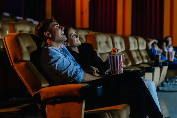 Ραντεβού το βράδυ στο σινεμά! Πορτρέτο ενός ευτυχισμένου νεαρού ζευγαριού που κάθεται στο αμφιθέατρο του κινηματογράφου τρώγοντας ποπ κορν και βλέποντας κωμωδία μαζί. Έννοια ψυχαγωγίας και ομαδικότητας - Φωτογραφία, εικόνα
