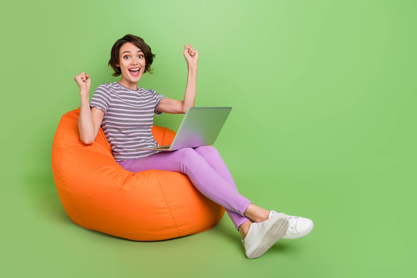 Photo of astonished funny young lady wear striped t-shirt sitting bean bag rising fists empty space modern device isolated green color background - Zdjęcie, obraz