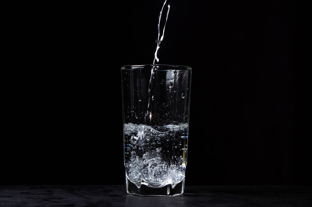 El agua mineral se vierte en un vaso sobre un fondo negro. Agua mineral potable útil. - Foto, imagen