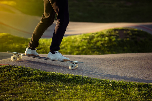 Gambe in pantaloni neri e scarpe da ginnastica bianche in sella al pattino, giovane pattinaggio a rotelle su uno skateboard, strada asfaltata e prato verde sullo sfondo - Foto, immagini