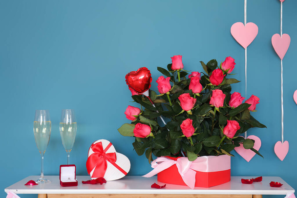 Schachteln mit Verlobungsring, Champagnergläsern und Rosen auf dem Tisch neben der blauen Wand im zum Valentinstag dekorierten Zimmer - Foto, Bild