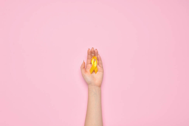 Mano femminile con nastro di consapevolezza dorata su sfondo rosa. Giornata internazionale del cancro infantile - Foto, immagini