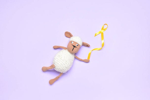 Golden awareness ribbon and toy sheep on violet background. International Childhood Cancer Day - Photo, Image
