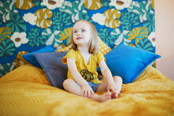 Adorable playful little girl sitting on bed in nicely decorated bedroom - Фото, изображение