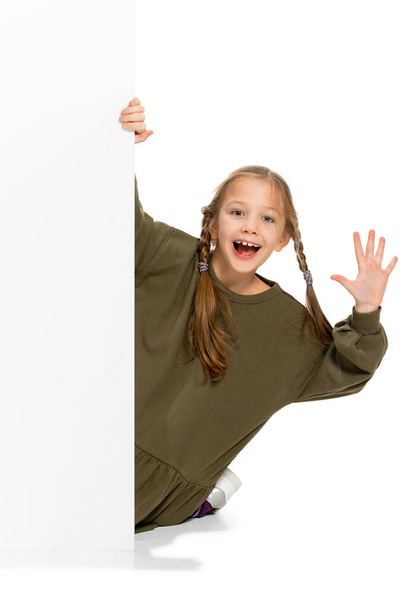 Lustiges Kind, kleines Mädchen, Schülerin in lässiger Kleidung, die hinter der Mauer isoliert auf weißem Hintergrund hervorlugt. Konzept von Kindheit, Emotionen, Studium - Foto, Bild