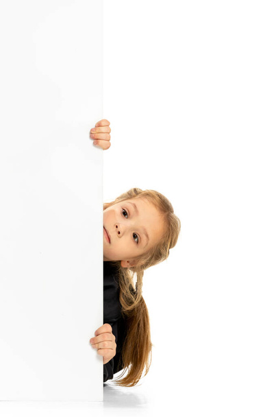 Funny child, little girl, pupil in casual style clothes peeking out from behind wall isolated on white background. Concept of childhood, emotions, study - Photo, image
