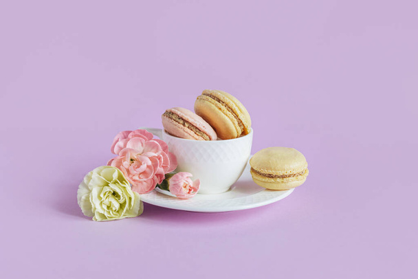 Вкусные французские макароны с цветами в белой чашке на фиолетовом фоне пастели. Место для текста. - Фото, изображение