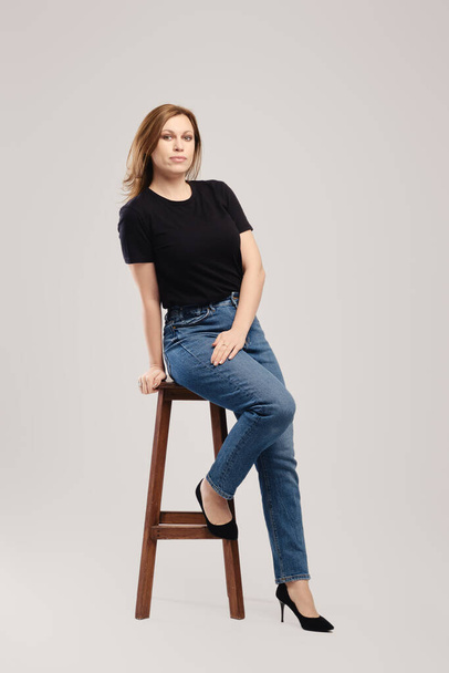 Young woman in black t shirt and jeans posing on tall wooden chair over grey background - Foto, Imagen