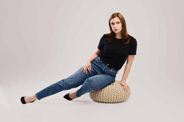 Young woman in casual black t shirt and jeans sits on the studio floor over grey background - Fotoğraf, Görsel