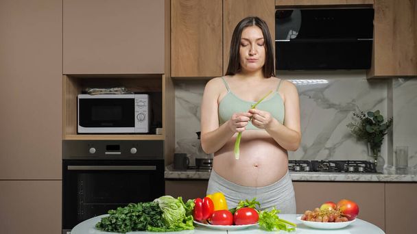 Pregnant woman peals and eats celery caressing bare belly - Zdjęcie, obraz
