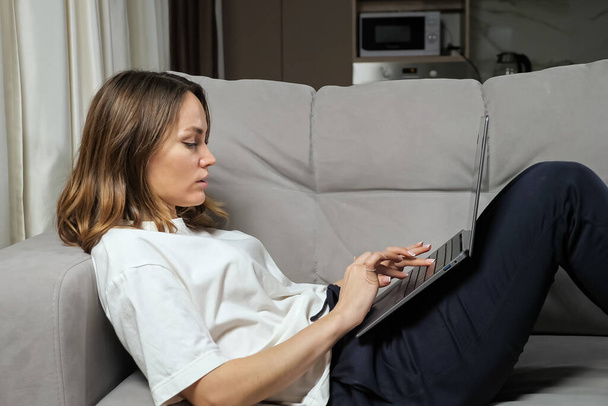 Freelancer woman works online via laptop lying on grey sofa - Photo, Image