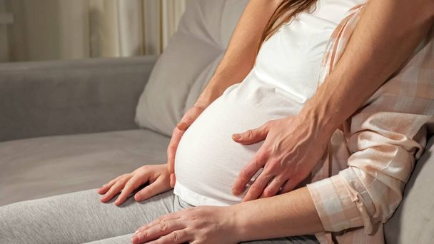 Young man caresses pregnant belly of wife sitting on sofa - Fotoğraf, Görsel