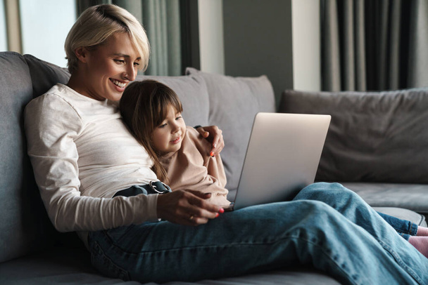 Happy young mother and her little daughter sitting on a couch with laptop computer, looking at screen, on a video call or watching movies - Фото, изображение