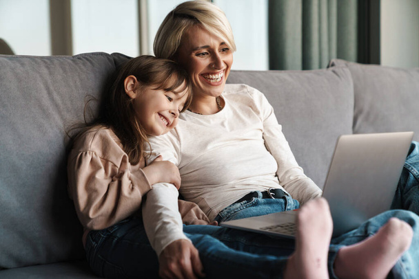 Happy young mother and her little daughter sitting on a couch with laptop computer, looking at screen, on a video call or watching movies - Φωτογραφία, εικόνα