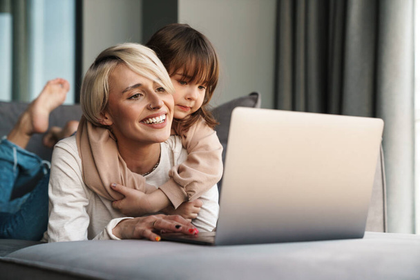 Happy young mother and her little daughter sitting on a couch with laptop computer, looking at screen, on a video call or watching movies - Foto, Imagen