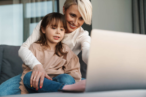 Happy young mother and her little daughter sitting on a couch with laptop computer, looking at screen, on a video call or watching movies - Foto, imagen