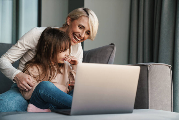 Happy young mother and her little daughter sitting on a couch with laptop computer, looking at screen, on a video call or watching movies - Fotoğraf, Görsel