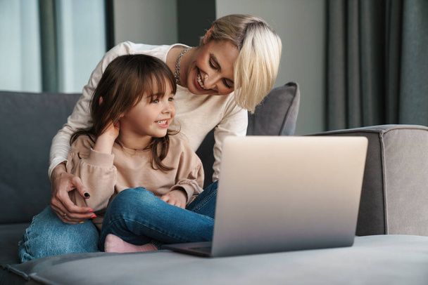 Happy young mother and her little daughter sitting on a couch with laptop computer, looking at screen, on a video call or watching movies - Foto, Bild