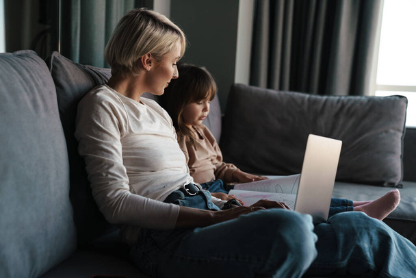 Onnellinen nuori äiti ja hänen pieni tyttärensä istuu sohvalla kannettavan tietokoneen kanssa, katselee näyttöä, videopuhelua tai katselee elokuvia - Valokuva, kuva