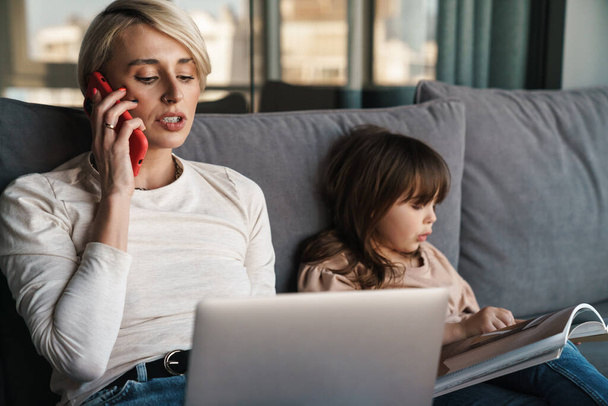 Happy young mother and her little daughter sitting on a couch with laptop computer, looking at screen, on a video call or watching movies, woman talking on mobile phone - Φωτογραφία, εικόνα