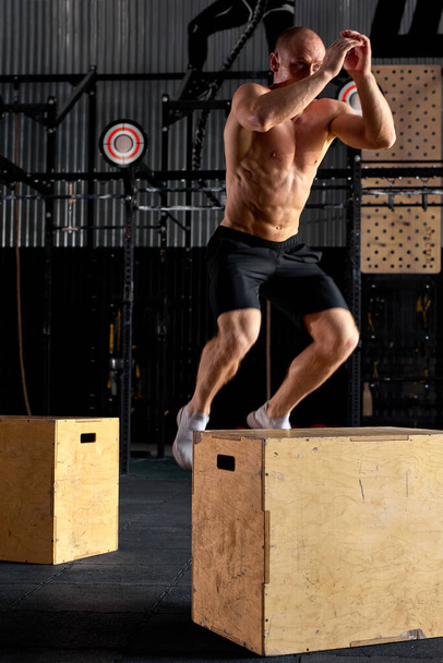 Young caucasian shirtless male is jumping on cross fit wooden box, at gym - Photo, image