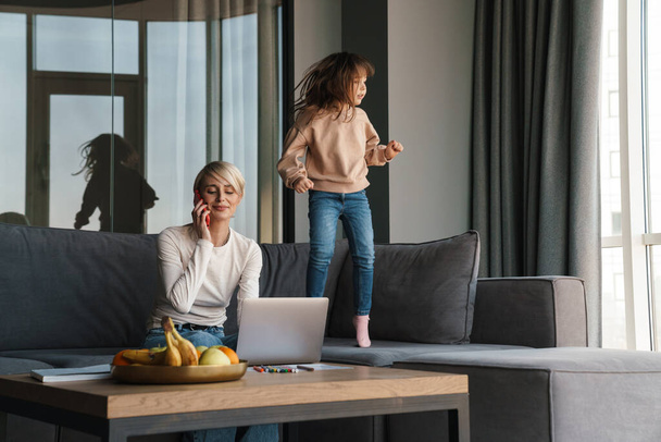 Happy young blonde mother working on laptop computer while her daughter jumping on a couch at home - Φωτογραφία, εικόνα