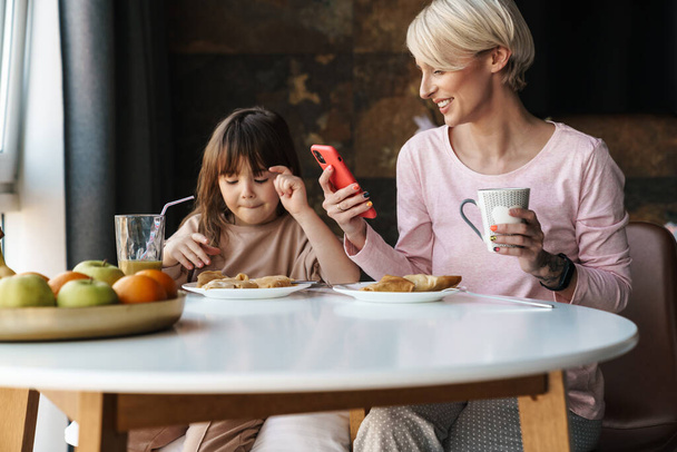 Boldog fiatal anya és kislánya együtt reggeliznek, anya mobiltelefont használ - Fotó, kép