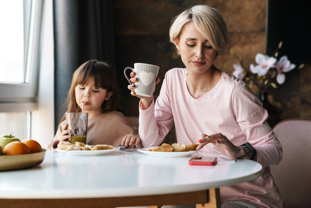 Blonde young mother and her little daughter having breakfast, mother using mobile phone - Фото, зображення