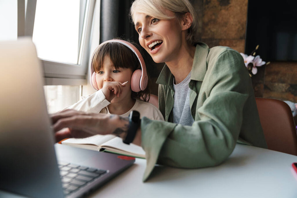 Boldog anya segít a lányának az iskolai házi feladatban, laptopot használ, tanul a karantén alatt, a kislány hallgatja a tanárt a fülhallgatóban - Fotó, kép