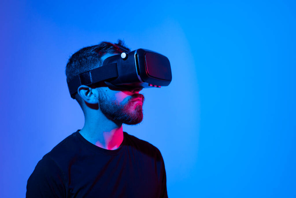 Virtual reality world. Bearded guy in vr headset, touching something while playing video game in metaverse with his friends, standing in neon light. - Photo, Image
