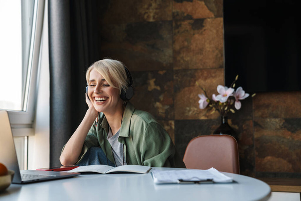 Smiling middle aged blonde woman with short hair studying or working on a video call via laptop computer while sitting at the table at home - 写真・画像