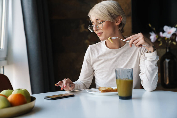 Sorrindo jovem mulher vestindo roupas casuais usando telefone celular enquanto sentado na mesa da cozinha tomando café da manhã - Foto, Imagem