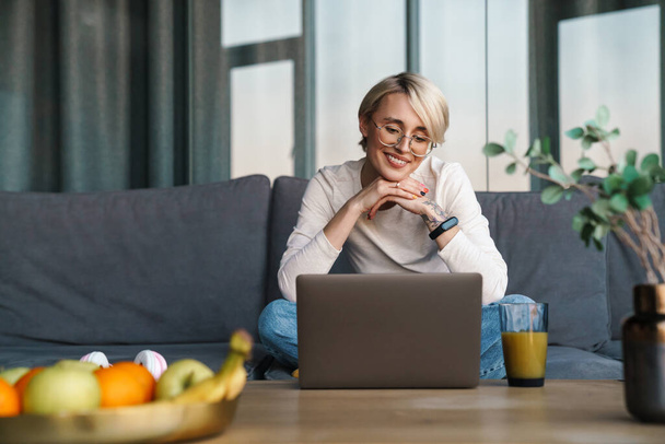Smiling mid aged blonde woman studying online via laptop computer in the living room - Photo, Image